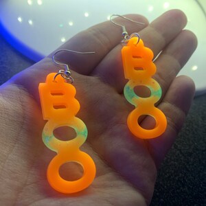 May include: A pair of Halloween-themed earrings spelling out "BOO" in bright orange, with a gradient of light green and glitter. The earrings are suspended from silver-colored fish hook ear wires.