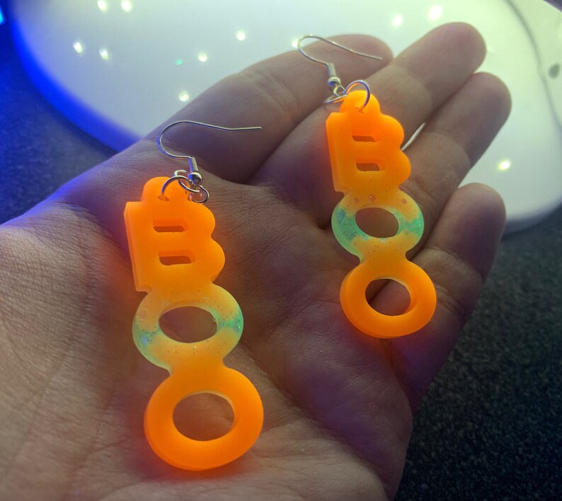 May include: A pair of Halloween-themed earrings spelling out "BOO" in bright orange, with a gradient of light green and glitter. The earrings are suspended from silver-colored fish hook ear wires.
