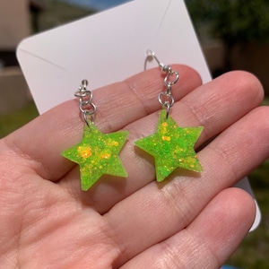 May include: A pair of neon green star-shaped earrings with silver-colored hooks. The stars are filled with gold glitter, creating a sparkling effect. The earrings are displayed against a soft background.