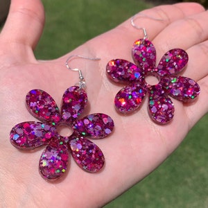 May include: A pair of pink flower-shaped earrings with iridescent glitter embedded in the resin. The earrings have a silver hook fastening.