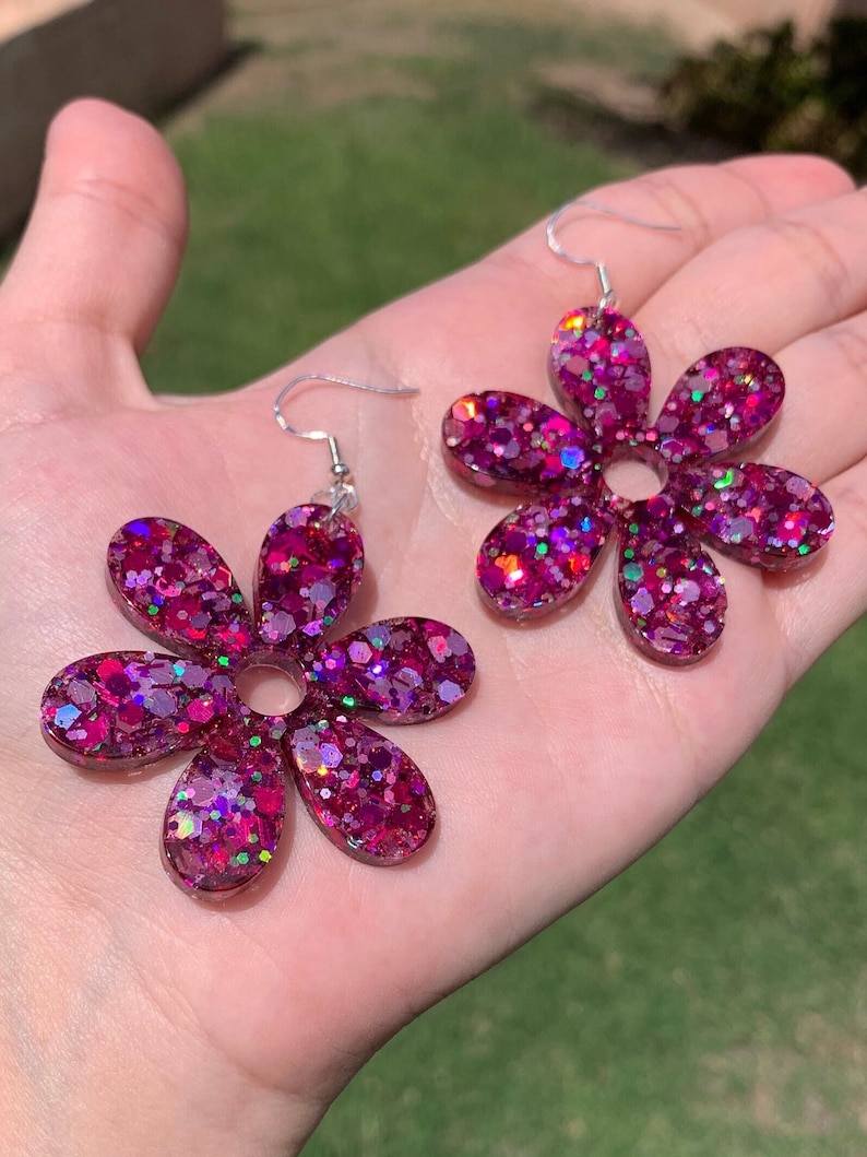 May include: A pair of pink flower-shaped earrings with iridescent glitter embedded in the resin. The earrings have a silver hook fastening.