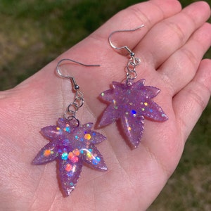 May include: A pair of silver dangle earrings with purple resin leaf charms. The leaves are covered in iridescent glitter.