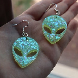 May include: A pair of green alien face earrings with iridescent glitter. The earrings are made of resin and have silver hooks.