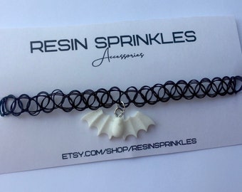 May include: A black choker necklace with a white resin bat charm. The charm is attached to the necklace with a silver chain. The necklace is on a white background with the text "RESIN SPRINKLES Accessories" and "ETSY.COM/SHOP/RESINSPRINKLES".