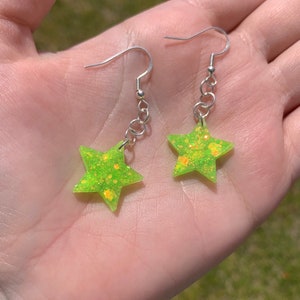 May include: A pair of star-shaped earrings. Each earring features a bright neon green star with orange glitter accents. The stars are suspended from silver-colored fish hook ear wires.
