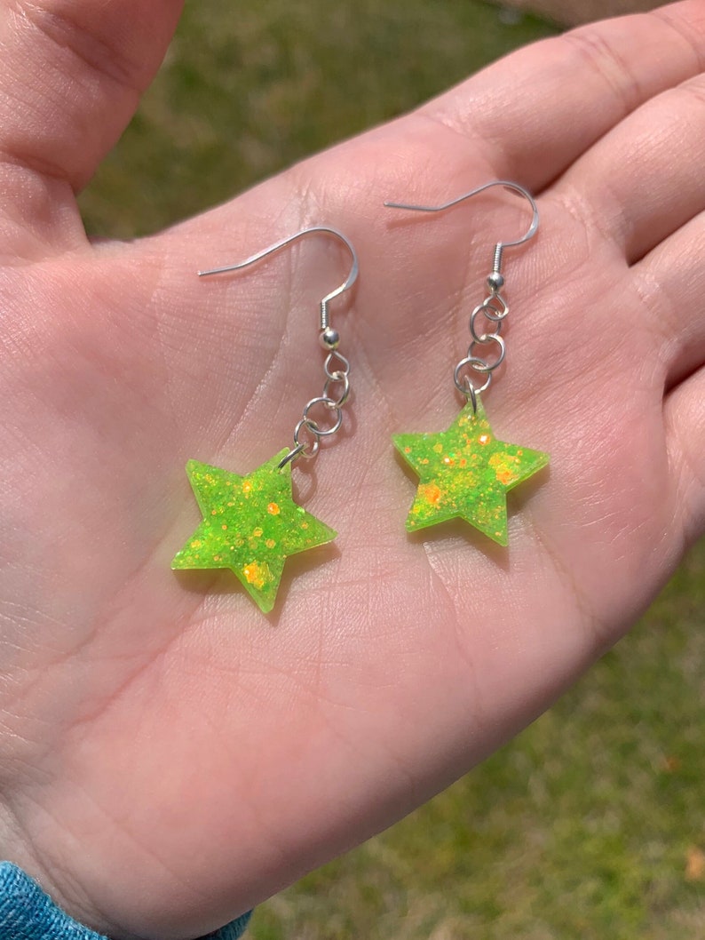 May include: A pair of star-shaped earrings. Each earring features a bright neon green star with orange glitter accents. The stars are suspended from silver-colored fish hook ear wires.