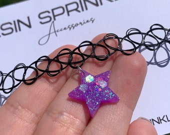 May include: A black choker necklace with a purple star charm. The star is made of resin and has glitter embedded in it. The star has a silver loop for attaching to the necklace.