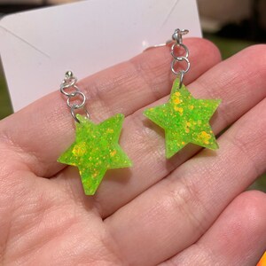 May include: A pair of neon green star-shaped earrings with silver-colored hooks. The earrings have a glittery texture and are displayed against a blurred background. The earrings are a fun and playful accessory.
