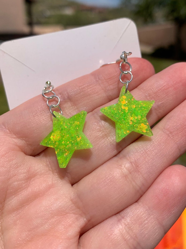 May include: A pair of neon green star-shaped earrings with silver-colored hooks. The earrings have a glittery texture and are displayed against a blurred background. The earrings are a fun and playful accessory.