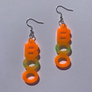 May include: A pair of orange acrylic earrings spelling out the word "BOO" in a vertical line. The earrings have silver hooks and a glittery green and yellow gradient effect.