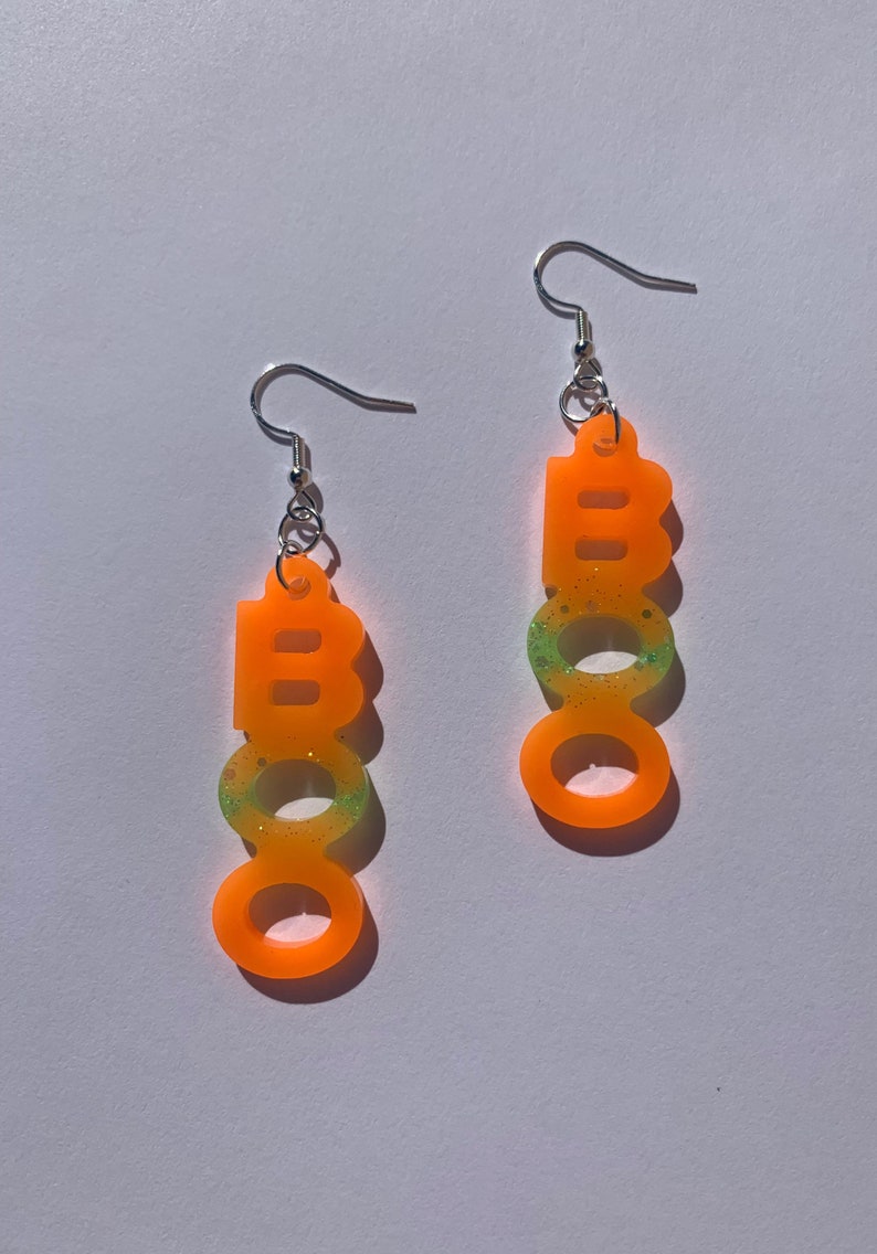 May include: A pair of orange acrylic earrings spelling out the word "BOO" in a vertical line. The earrings have silver hooks and a glittery green and yellow gradient effect.