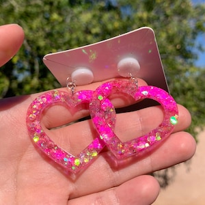 May include: A pair of pink heart-shaped earrings with iridescent glitter embedded in the resin. The earrings are on a white card.