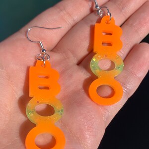 May include: Orange "BOO" earrings with silver hooks. The earrings have a gradient of yellow and green glitter at the bottom of the "O"s. These Halloween-themed earrings are a fun accessory for the spooky season.