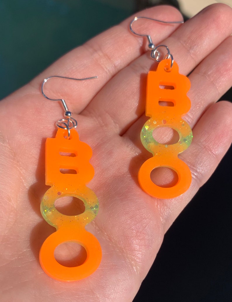 May include: Orange "BOO" earrings with silver hooks. The earrings have a gradient of yellow and green glitter at the bottom of the "O"s. These Halloween-themed earrings are a fun accessory for the spooky season.