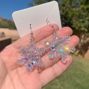 May include: A pair of snowflake-shaped earrings. The earrings are clear with iridescent glitter and small, colorful specks. They hang from silver-colored fish hook ear wires. The earrings are held in a hand against a background of green grass and a blue sky.