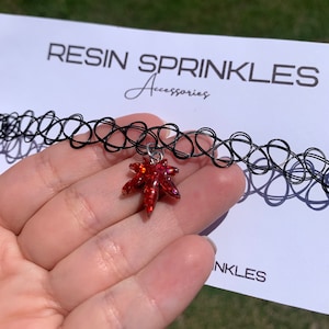 May include: Black choker necklace with a red glitter leaf pendant. The pendant is a five-pointed leaf shape with a shiny, textured surface. The text "RESIN SPRINKLES Accessories" is printed on a white card in the background.
