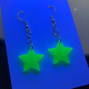May include: A pair of dangling earrings featuring bright neon green star-shaped pendants. The stars are suspended from silver-colored fish hook ear wires and chain links. The earrings are displayed on a blue background.