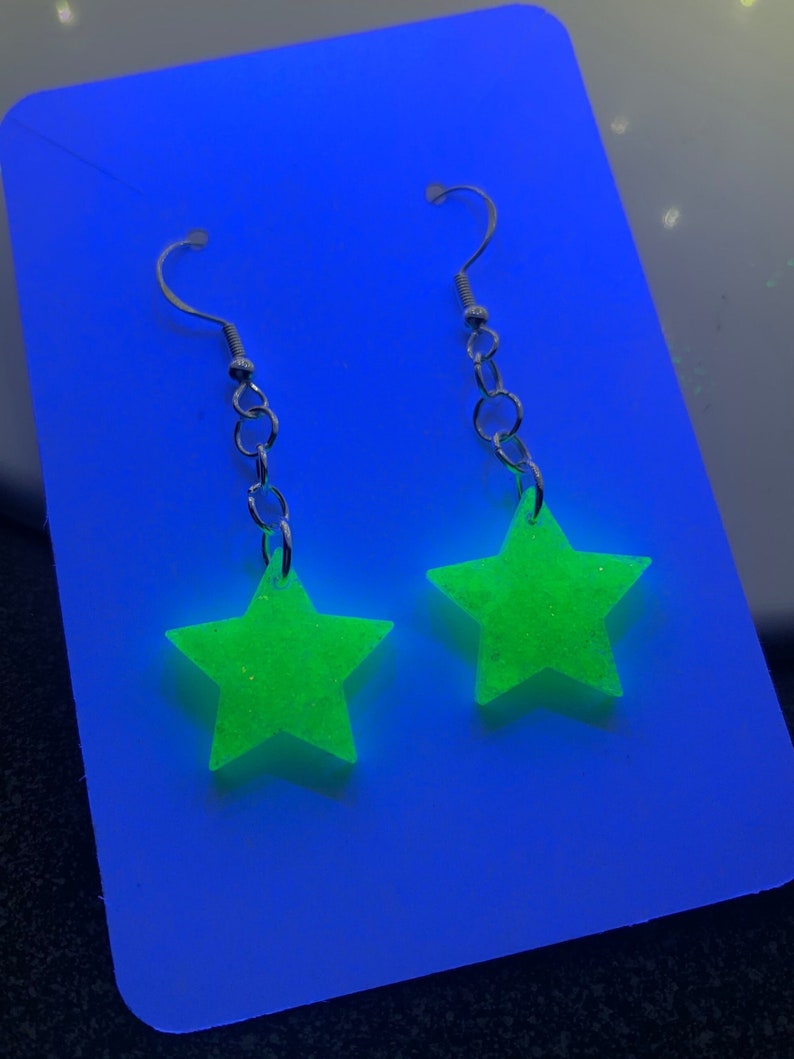 May include: A pair of dangling earrings featuring bright neon green star-shaped pendants. The stars are suspended from silver-colored fish hook ear wires and chain links. The earrings are displayed on a blue background.