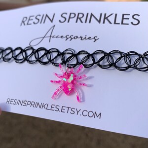 May include: A pink glitter spider charm on a black choker necklace. The necklace is made of elastic and has a clasp closure. The charm is attached to the necklace with a silver ring. The image also includes the text "RESINSPRINKLES.ETSY.COM".