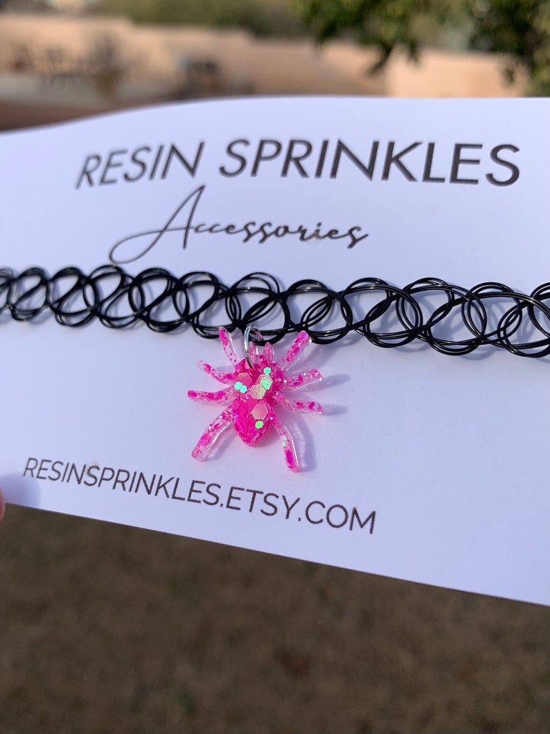 May include: A pink glitter spider charm on a black choker necklace. The necklace is made of elastic and has a clasp closure. The charm is attached to the necklace with a silver ring. The image also includes the text "RESINSPRINKLES.ETSY.COM".