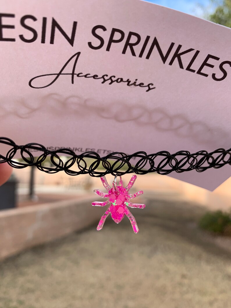 May include: A black choker necklace with a pink glitter spider charm. The charm is made of resin and has a sparkly finish. The choker is made of elastic and is adjustable.