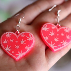 May include: A pair of pink heart-shaped earrings with a white marijuana leaf pattern.