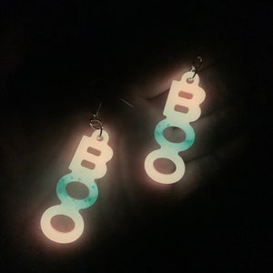 May include: A pair of dangling earrings spelling out "BOO" in a vertical arrangement. The earrings are white with a teal and blue speckled design, and glow in the dark. The earrings are suspended from silver-colored hooks.