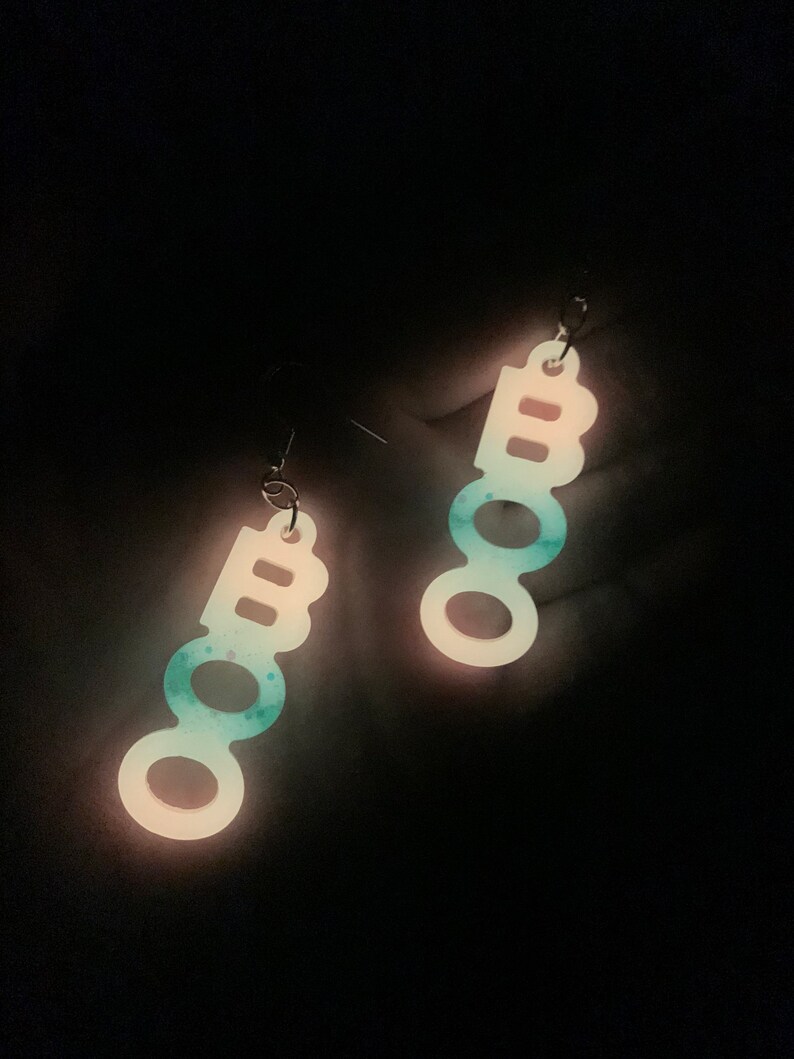 May include: A pair of dangling earrings spelling out "BOO" in a vertical arrangement. The earrings are white with a teal and blue speckled design, and glow in the dark. The earrings are suspended from silver-colored hooks.