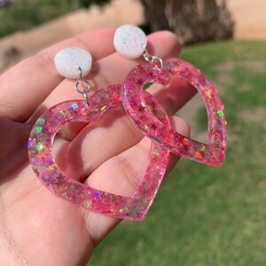 May include: Pink heart-shaped earrings with glitter and iridescent stars. The earrings have a white, sparkly, round charm at the top.