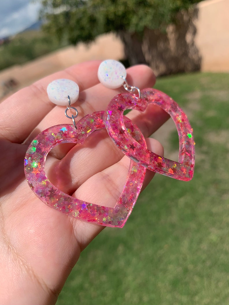 May include: Pink heart-shaped earrings with glitter and iridescent stars. The earrings have a white, sparkly, round charm at the top.