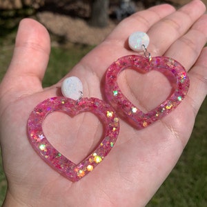 May include: A pair of pink heart-shaped earrings with iridescent glitter and white opal-like accents.