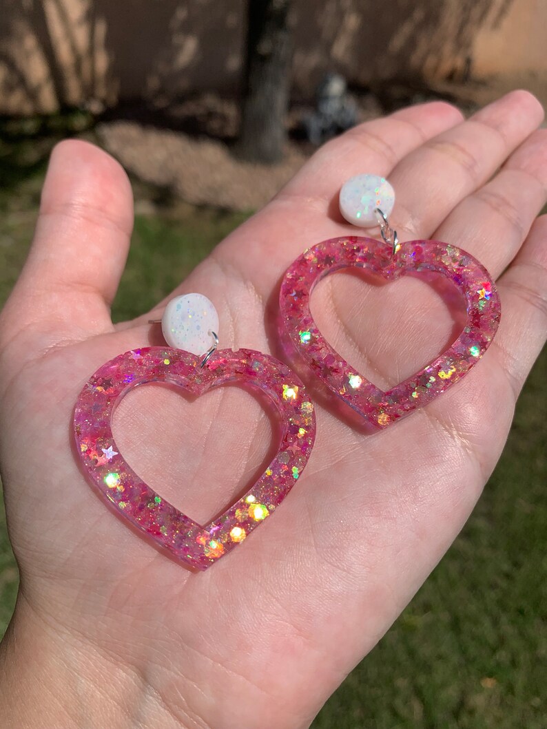 May include: A pair of pink heart-shaped earrings with iridescent glitter and white opal-like accents.