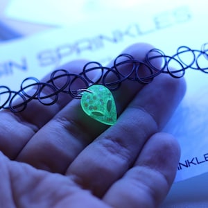 May include: Black tattoo choker necklace with a glowing green alien pendant. The alien pendant is filled with glitter and has a silver bail. The necklace is held in a hand against a blue background.