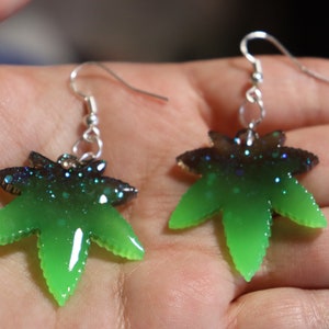 May include: A pair of silver dangle earrings with green and black resin marijuana leaf charms. The leaves have a glittery finish.