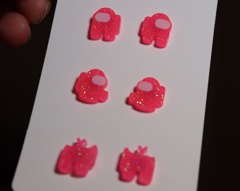 May include: A set of four pink glitter earrings shaped like the characters from the video game Among Us. The earrings are a light pink color with white accents and glitter throughout.