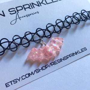 May include: A black choker necklace with a pink glitter bat charm. The charm is attached to the choker with a silver chain. The choker is on a white background with the text "N SPRINKLES Accessories" and "ETSY.COM/SHOP/RESINSPRINKLES".