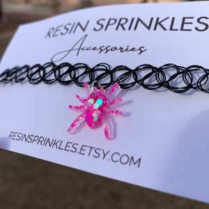 May include: A black choker necklace with a pink glitter spider charm. The charm is made of resin and has a silver ring for attaching it to the necklace. The necklace is on a white card with the text "RESIN SPRINKLES Accessories" and "RESINSPRINKLES.ETSY.COM".