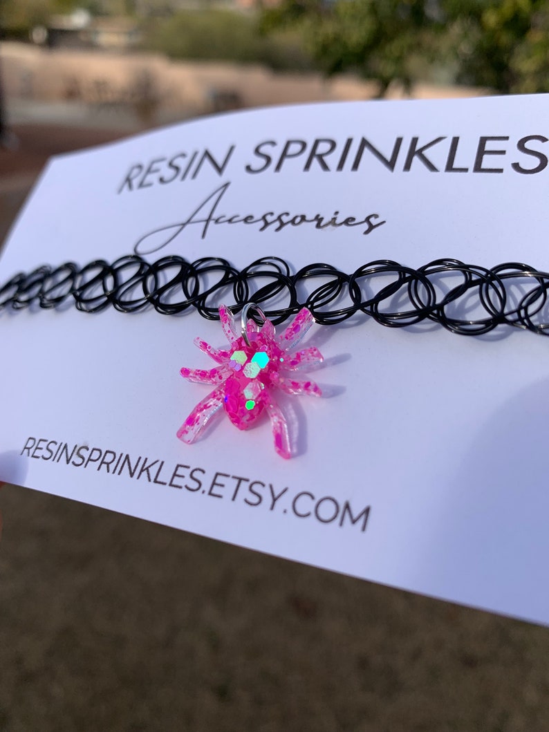 May include: A black choker necklace with a pink glitter spider charm. The charm is made of resin and has a silver ring for attaching it to the necklace. The necklace is on a white card with the text "RESIN SPRINKLES Accessories" and "RESINSPRINKLES.ETSY.COM".