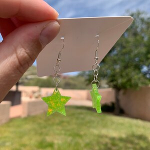 May include: A pair of star-shaped earrings. The earrings are lime green with yellow speckles and hang from silver-colored hooks. The earrings are displayed against a white card, with a blurred outdoor background of green grass and a blue sky.