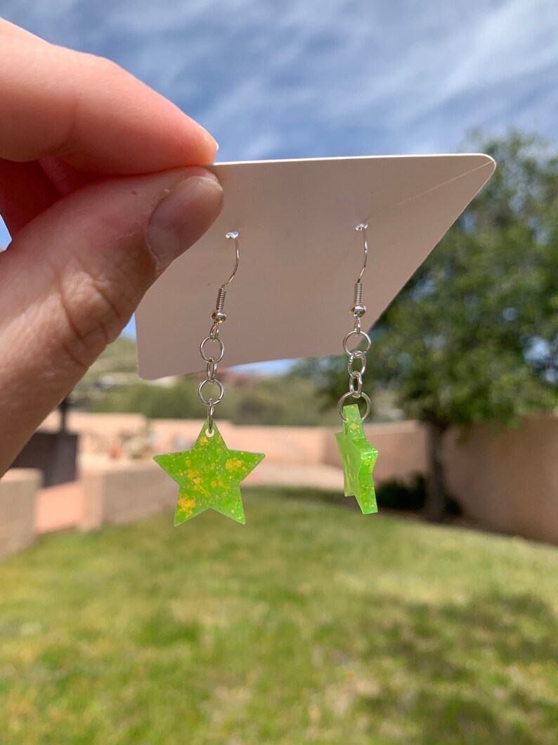 May include: A pair of star-shaped earrings. The earrings are lime green with yellow speckles and hang from silver-colored hooks. The earrings are displayed against a white card, with a blurred outdoor background of green grass and a blue sky.