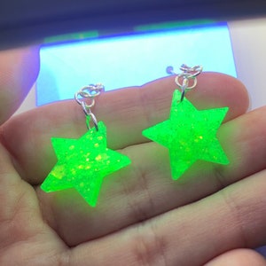 May include: A pair of neon green star-shaped earrings with silver-colored hooks. The earrings have a glittery texture and are held in a person's hand. The background is out of focus.
