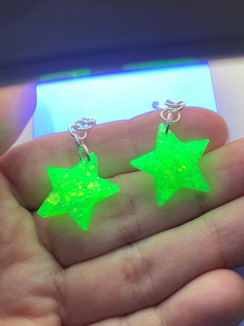 May include: A pair of neon green star-shaped earrings with silver-colored hooks. The earrings have a glittery texture and are held in a person's hand. The background is out of focus.