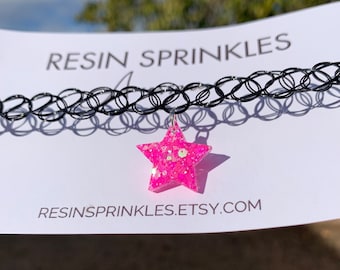 May include: A black choker necklace with a pink glitter star charm. The charm is made of resin and has a silver loop for attaching to the necklace. The text "RESINSPRINKLES.ETSY.COM" is printed on a white card behind the necklace.