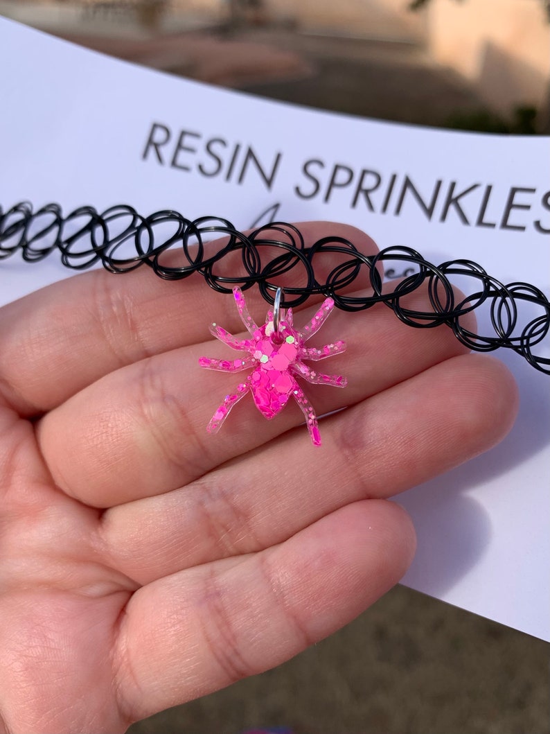May include: A black choker necklace with a pink glitter spider charm. The charm is made of resin and has a silver loop for attaching to the necklace. The text "RESIN SPRINKLES" is visible in the background.