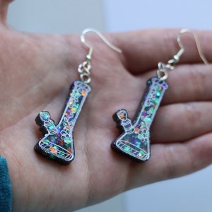 May include: A pair of dangle earrings featuring a black resin bong with iridescent glitter and white floral details. The earrings have silver hooks.