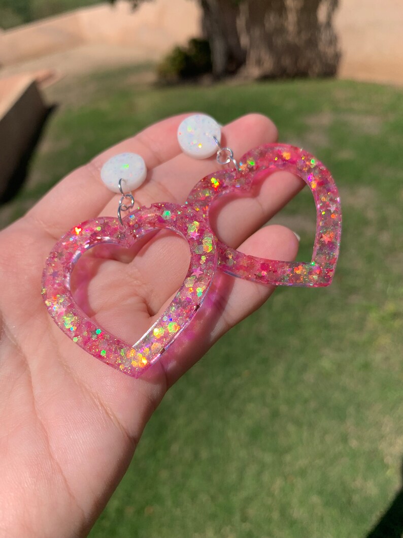 May include: A pair of pink heart-shaped earrings with iridescent glitter and white round accents.