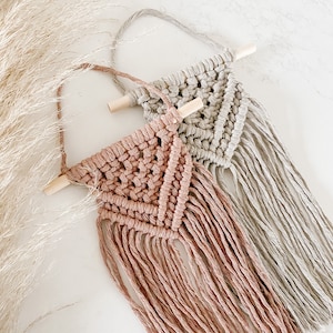 May include: Two macrame wall hangings with wooden dowels. One is a light pink colour and the other is a light beige colour.
