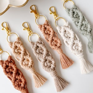 May include: Six handmade macrame keychains in various shades of brown, beige, and green. Each keychain features a tassel and a gold metal key ring.
