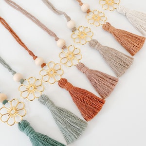 May include: Seven decorative tassels with gold flower charms and wooden beads. The tassels are in various shades of brown, beige, and green.