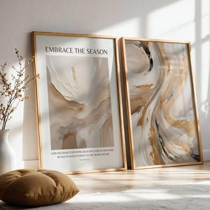 May include: Two framed art prints with abstract designs in beige, brown, and gold. One print reads "EMBRACE THE SEASON" and the other reads "NOTHING CAN SEPARATE US FROM GOD'S LOVE". A white vase with branches and a brown cushion are in the foreground.